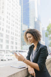 Female entrepreneur using smart phone while leaning on retaining wall in city - BOYF01942