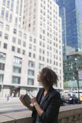 Thoughtful Afro businesswoman with diary looking at office buildings in city - BOYF01957