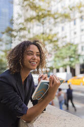 Lächelnde junge Geschäftsfrau mit Tagebuch in der Hand, während sie sich auf eine Stützmauer in der Stadt stützt - BOYF01963