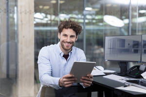 Lächelnder Geschäftsmann, der ein digitales Tablet hält, während er in der Nähe des Schreibtischs im Büro sitzt - FKF04023