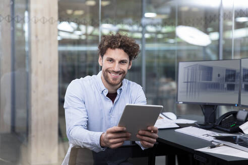Smiling businessman holding digital tablet while sitting near desk at office - FKF04023
