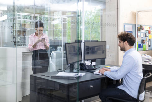 Junger männlicher Unternehmer bei der Arbeit, während eine Kollegin im Büro ein Smartphone benutzt - FKF04029