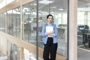 Geschäftsfrau, die ein Dokument hält, während sie gegen eine Glaswand im Büro steht - FKF04038