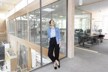 Thoughtful businesswoman leaning on glass wall in factory - FKF04042