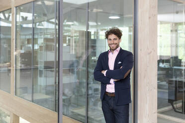 Smiling businessman leaning on glass wall at office - FKF04043