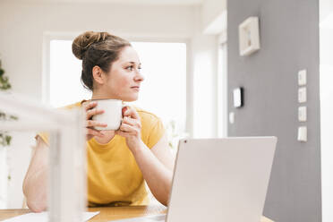 Unternehmerin hält Kaffeetasse, während sie am Schreibtisch sitzt und wegschaut - UUF22901