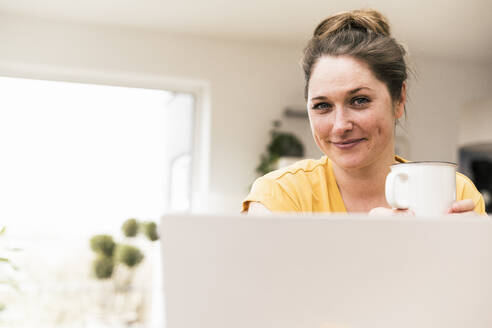 Smiling businesswoman holding coffee cup while taking break from work at home - UUF22903