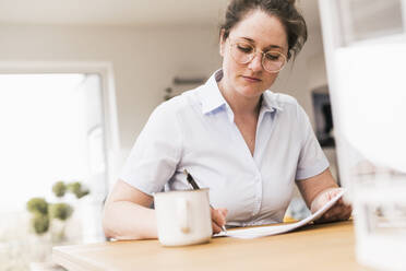 Female professional wearing eyeglasses writing on document while sitting at desk - UUF22922