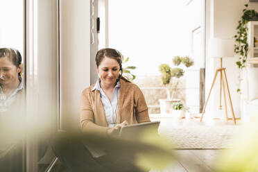 Smiling female entrepreneur wearing headphones using digital tablet - UUF22950