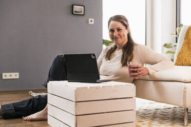 Smiling woman with glass of juice and digital tablet sitting in living room - UUF22955