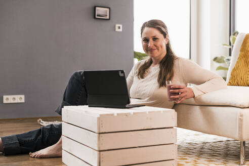 Lächelnde Frau mit einem Glas Saft und einem digitalen Tablet im Wohnzimmer sitzend - UUF22955