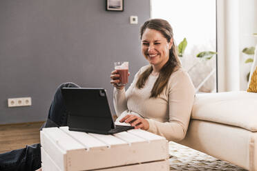 Fröhliche Frau trinkt Saft und sitzt mit einem Tablet im Wohnzimmer - UUF22957