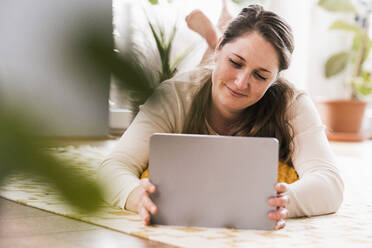 Mid adult woman using digital tablet while lying on carpet in living room - UUF22958