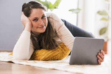 Entspannte Frau mit digitalem Tablet, die zu Hause auf dem Teppich liegt - UUF22959