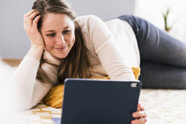 Lächelnde Frau sieht sich ein Video auf einem Tablet an, während sie auf dem Teppich im Wohnzimmer liegt - UUF22960