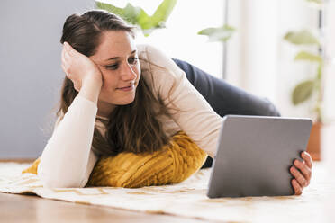 Entspannte Frau, die zu Hause auf dem Teppich liegend ein Video über ihr Tablet ansieht - UUF22961