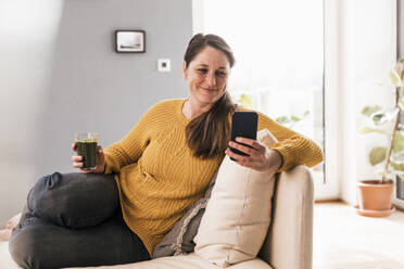 Smiling woman with glass of juice using smart phone sitting on sofa in living room - UUF22964