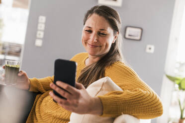 Glückliche erwachsene Frau, die ein Mobiltelefon im Wohnzimmer benutzt - UUF22965