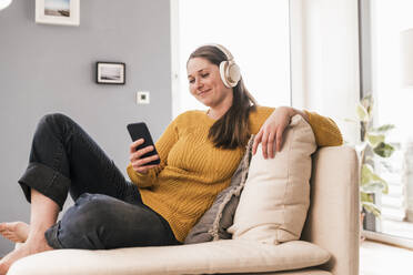 Lächelnde Frau mittleren Alters, die zu Hause Musik über Kopfhörer hört - UUF22968