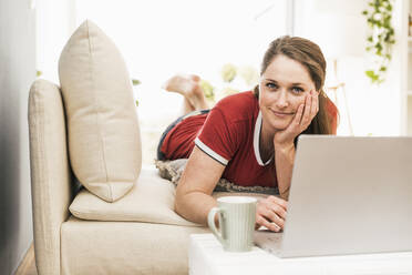 Mid adult woman with laptop lying on sofa in living room - UUF22971