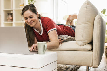 Mittlere erwachsene Frau, die einen Laptop benutzt, während sie auf dem Sofa im Wohnzimmer liegt - UUF22973