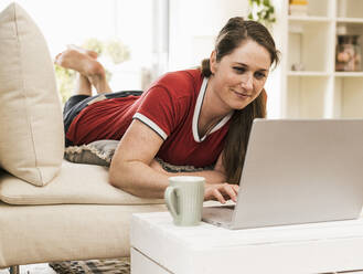 Frau benutzt Laptop, während sie zu Hause auf dem Sofa liegt - UUF22974