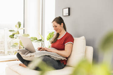 Woman sitting on sofa using laptop at home - UUF22975