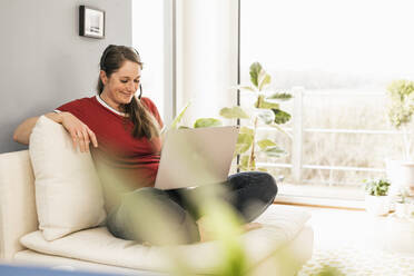 Smiling woman wearing headphones sitting with laptop on sofa in living room - UUF22976