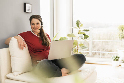 Lächelnde Frau mit Laptop und Kopfhörern, die auf dem Sofa im Wohnzimmer sitzt - UUF22978