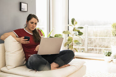 Frau mit Smartphone und Laptop im Wohnzimmer - UUF22979