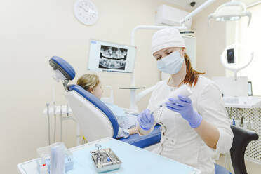 Female dentist wearing protective face mask preparing syringe for injection with little girl sitting in background - EYAF01541