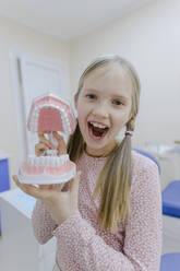 Portrait of little blonde girl standing in dentists office with dentures in hands - EYAF01545