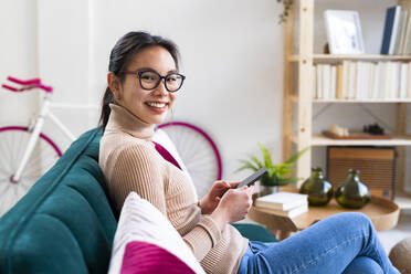 Junge Frau mit Brille sitzt mit Handy zu Hause - GIOF11651