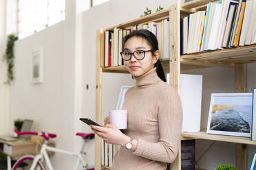 Woman with coffee cup and mobile phone standing by bookshelf at home - GIOF11658