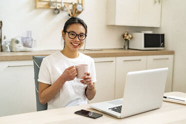 Lächelnde junge Frau sitzt mit Kaffeetasse und Laptop zu Hause - GIOF11665