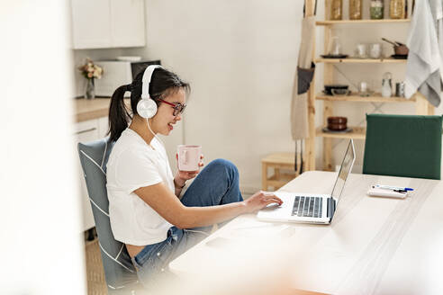 Frau mit Kopfhörern und Kaffeetasse, die zu Hause einen Laptop benutzt - GIOF11675
