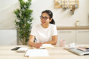 Young woman smiling while sitting by table at home - GIOF11676