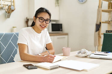 Smiling woman wearing eyeglasses sitting by book at home - GIOF11678