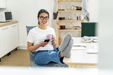 Junge Frau mit Kaffeetasse und Mobiltelefon, lächelnd zu Hause sitzend - GIOF11683