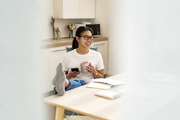 Young woman with coffee cup and mobile phone leaning legs on table at home - GIOF11684