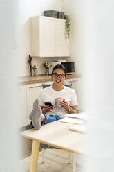 Woman with mobile phone and coffee cup smiling while relaxing by table at home - GIOF11685