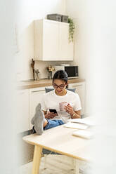 Lächelnde junge Frau, die ein Mobiltelefon benutzt, während sie zu Hause bei einem Kaffee sitzt - GIOF11686