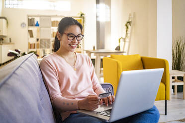 Lächelnde junge Frau, die eine Kreditkarte in der Hand hält, während sie zu Hause am Laptop sitzt - GIOF11707