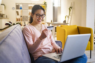 Happy woman sitting with credit card and laptop at home - GIOF11708