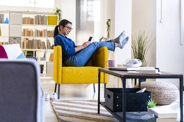 Woman using mobile phone while sitting on chair at home - GIOF11716