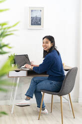 Young woman sitting with laptop by table at home office - GIOF11724