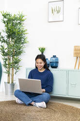Lächelnde Frau, die einen Laptop benutzt, während sie zu Hause auf einem Juteteppich sitzt - GIOF11727