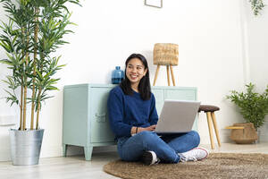 Lächelnde junge Frau sitzt mit Laptop auf einem Juteteppich zu Hause - GIOF11729