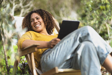 Smiling woman with digital tablet looking away while sitting on deck chair in vegetable garden - SBOF03092