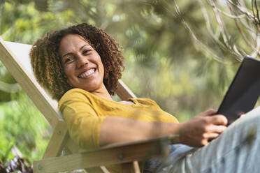 Smiling curly haired woman holding digital tablet while sitting on chair in garden - SBOF03093